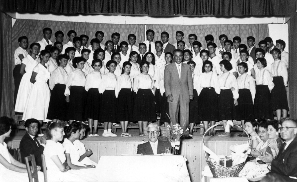 ՄԿՀ-ի երգչախումբը 1957-ի ամավերջի հանդէսին օրը Խմբավար պրն. Վ. Պէտէլեան բեմին վրայ Բեմէն վար, կեդրոնը նստած` տնօրէն Բիւզանդ Եղիայեան